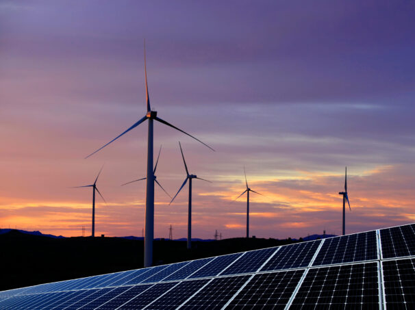 Solar photovoltaic panels and wind turbines in front of a colorful sunset.